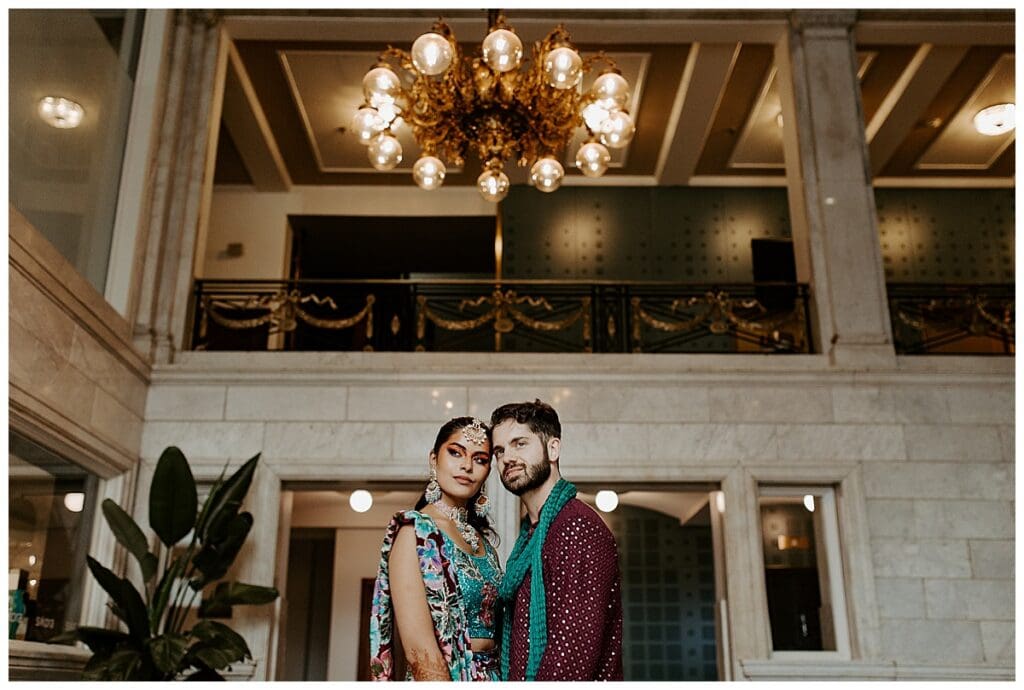 Aarushi and Shane standing together under a chandelier in the grand interior of the Lumber Exchange in Minneapolis